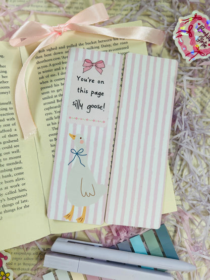 Set of three pastel bookmarks with silly goose and bow designs, placed on an open book with soft pink ribbon, shredded filler, and pens.