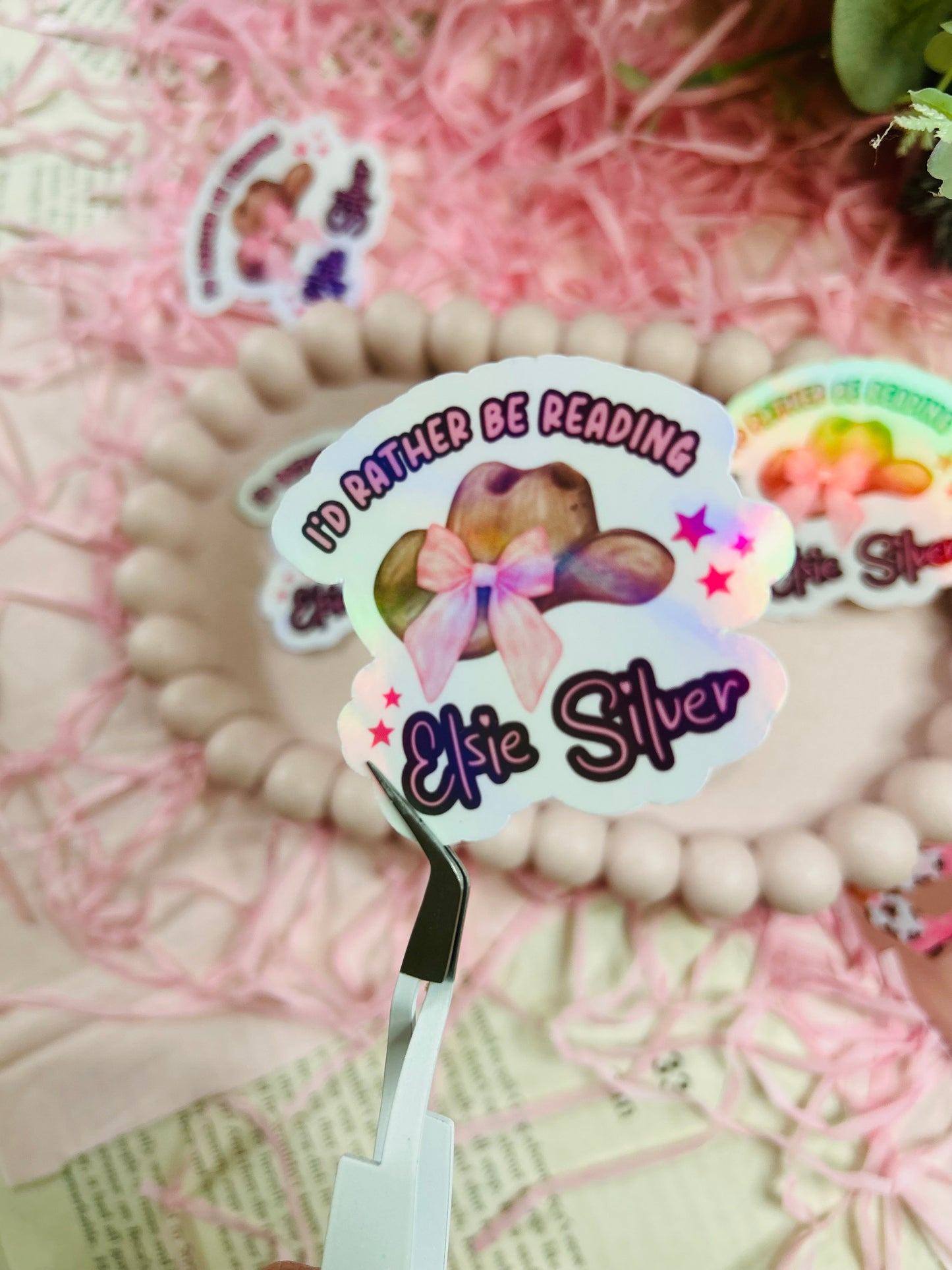 Holographic sticker with a pink-bowed cowboy hat and text reading "I'd Rather Be Reading Elsie Silver" on a bookish background with pink shredded paper