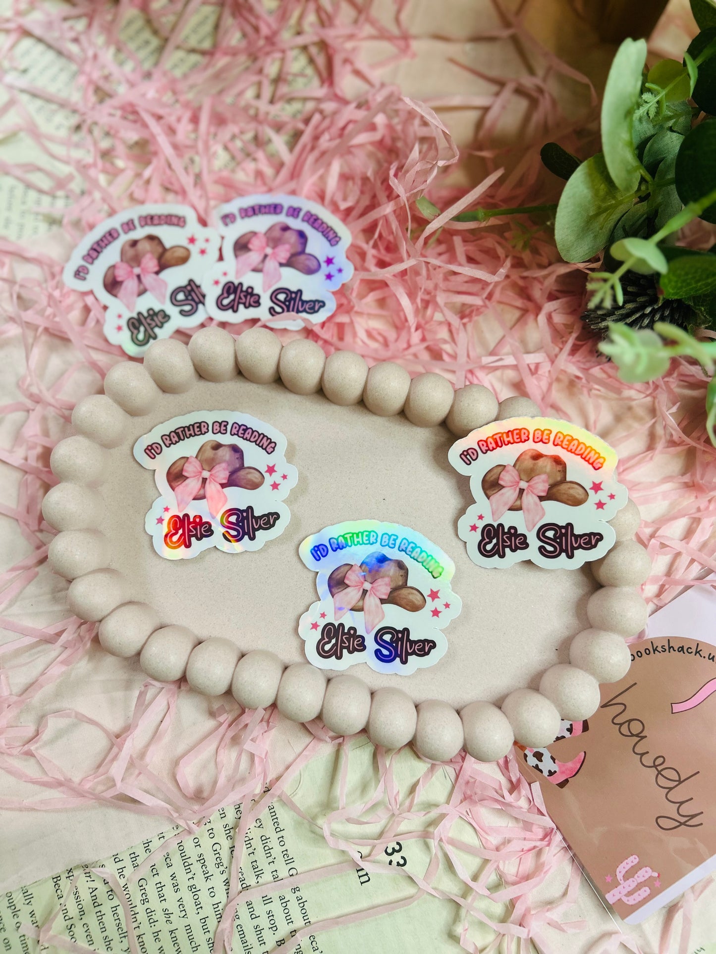 Holographic sticker with a pink-bowed cowboy hat and text reading "I'd Rather Be Reading Elsie Silver" on a bookish background with pink shredded paper