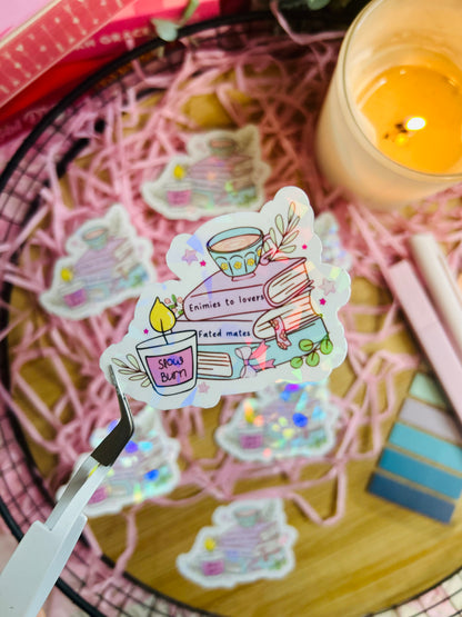 Holographic book stack sticker featuring a tea cup, books labelled with romance tropes (Enemies to Lovers, Slow Burn, Fated Mates), and cosy bookish details.
