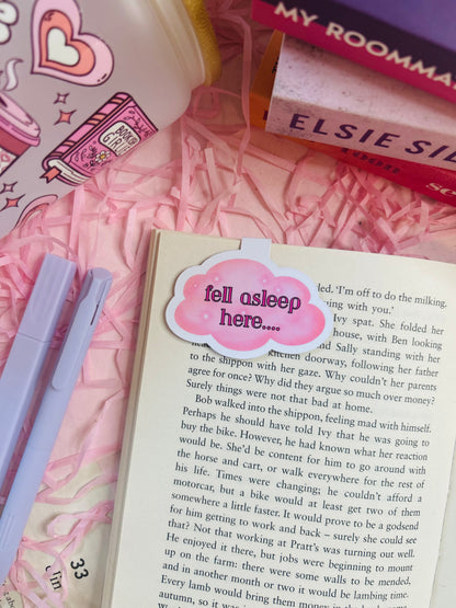 Cloud-shaped magnetic bookmark with the phrase ‘Fell Asleep Here...’ on soft pastel packaging, styled on an open book with cosy pink surroundings.
