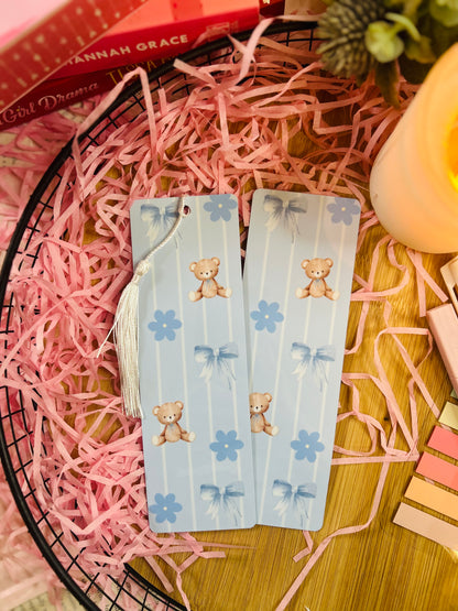 A pair of blue teddy bear-themed bookmarks with pastel vertical stripes, delicate bows, and floral accents. One bookmark features a white tassel, while the other remains plain.