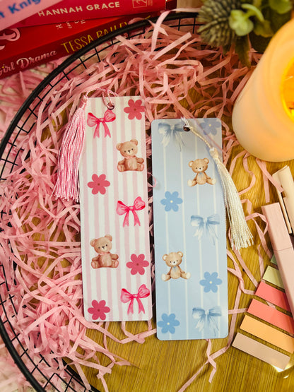 A set of two teddy bear-themed bookmarks, one in pink and one in blue, featuring soft pastel stripes, bows, and floral accents. Each bookmark has a matching tassel