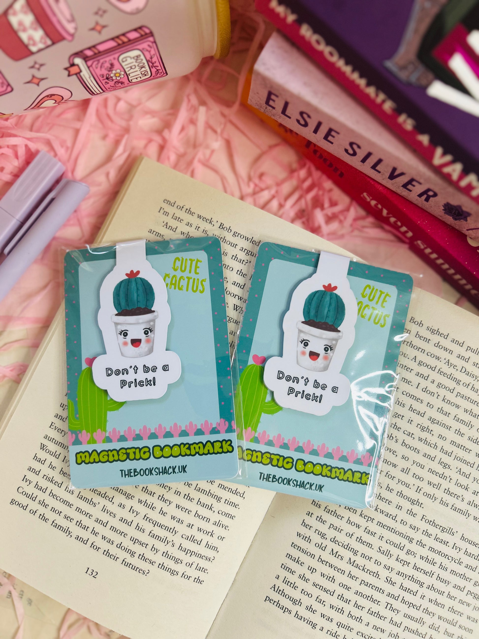 Funny cactus magnetic bookmark with the phrase ‘Don’t Be a Prick’ placed on an open book with pink shredded filler, pens, and books nearby.