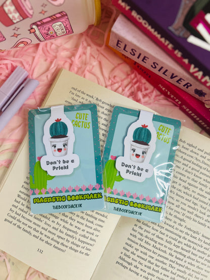 Funny cactus magnetic bookmark with the phrase ‘Don’t Be a Prick’ placed on an open book with pink shredded filler, pens, and books nearby.