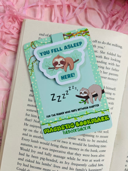 Sloth magnetic bookmark that says ‘You Fell Asleep Here’ placed on an open book with bookish stickers, pens, and novels in the background.