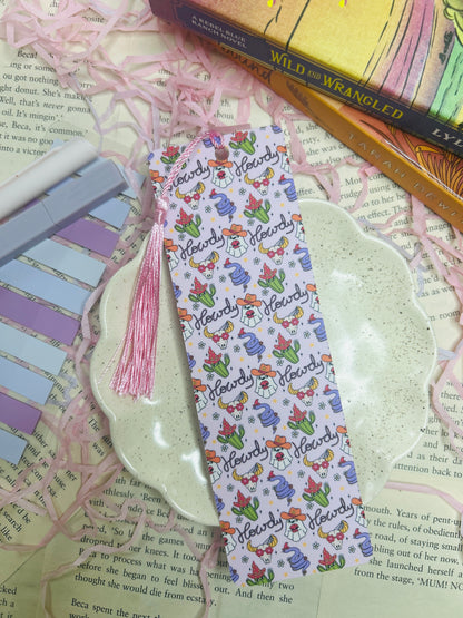Double-sided western bookmark with repeating “Howdy” design featuring cowboy hats, boots, flowers, cacti, and cow-print accents, finished with a pink tassel.