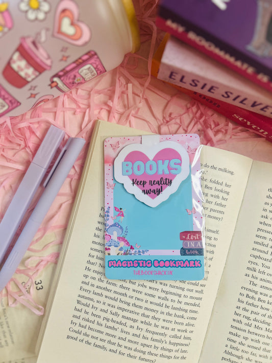 Heart-shaped pink magnetic bookmark with the phrase ‘Keep Reality Away’ on an open book surrounded by pink bookish accessories.