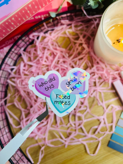Holographic book trope sticker featuring pastel candy hearts with the phrases "Fated Mates," "Slow Burn," and "Who Did This?" held by tweezers over pink shredded paper.