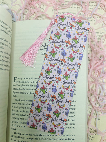 Double-sided western bookmark with repeating “Howdy” design featuring cowboy hats, boots, flowers, cacti, and cow-print accents, finished with a pink tassel.