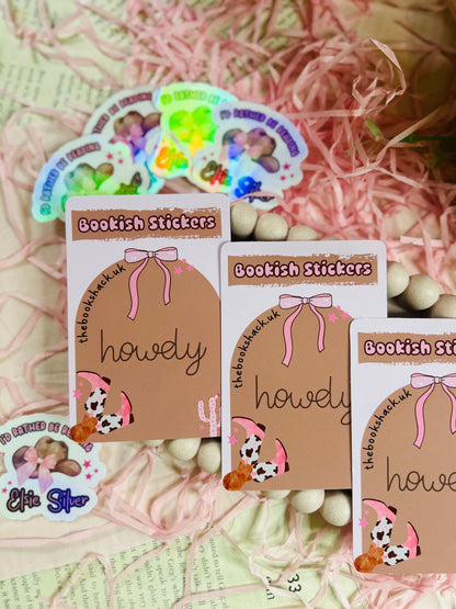 Holographic sticker with a pink-bowed cowboy hat and text reading "I'd Rather Be Reading Elsie Silver" on a bookish background with pink shredded paper