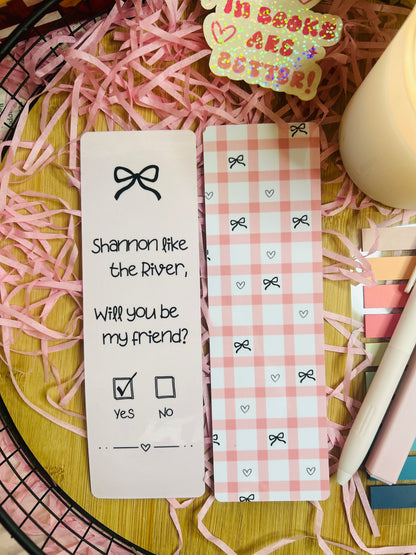 Two pink gingham bookmarks inspired by Binding 13, featuring a friendship note design with the phrase “Shannon like the River, Will you be my friend?” on one and a pink bow pattern on the other, styled with soft bookish decor.