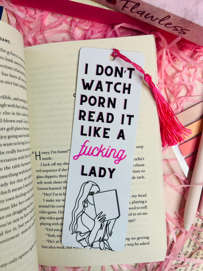 A sassy tassel bookmark with the quote 'I Don’t Watch, I Read It Like a F***ing Lady,' placed on a romance book, surrounded by pink shredded paper, pens, and other books