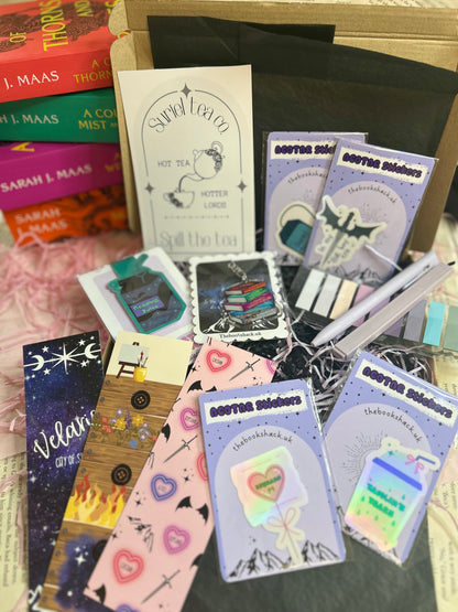 Flat lay of an ACOTAR-themed mystery bookish bundle featuring double-sided bookmarks, holographic stickers, a keyring, art print, annotate tabs, pen and highlighter. Items are displayed with Sarah J. Maas books and pink shredded paper, creating a cosy, fantasy-inspired aesthetic.