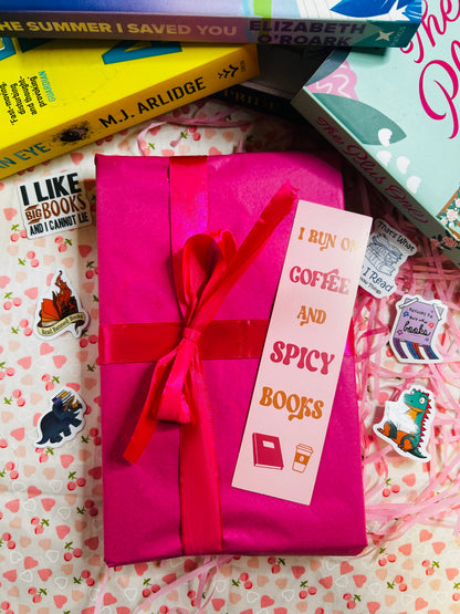  A pink bookish gift box wrapped with a red ribbon, featuring a bookmark that says "I run on coffee and spicy books." 