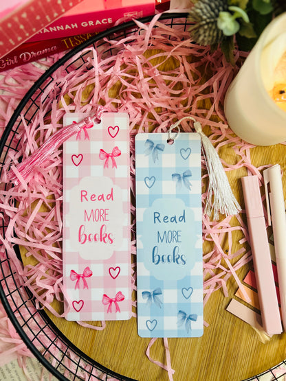 A set of pink and blue gingham bookmarks with delicate bow designs and the phrase "Read More Books," each featuring a matching tassel, surrounded by pink shredded paper, books, and cosy reading accessories.
