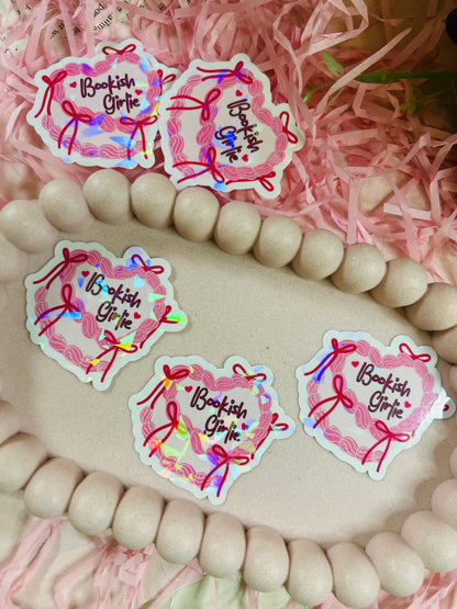 Holographic “Bookish Girlie” vinyl sticker with pink bows and rainbow shimmer, displayed with pink shredded paper and neutral wooden bead accents – perfect for aesthetic book lovers.