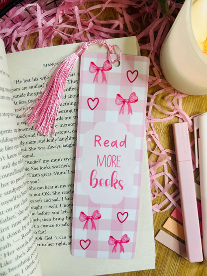 A pink gingham bookmark with delicate bow and heart designs, featuring the phrase "Read More Books" in red lettering. The bookmark is adorned with a soft pink tassel and placed inside an open book, surrounded by pink shredded paper, a candle, and pastel stationery for a cosy reading aesthetic.