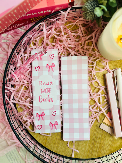 A pink gingham bookmark with delicate bow and heart designs, featuring the phrase "Read More Books" in red lettering. The bookmark is adorned with a soft pink tassel and placed inside an open book, surrounded by pink shredded paper, a candle, and pastel stationery for a cosy reading aesthetic.