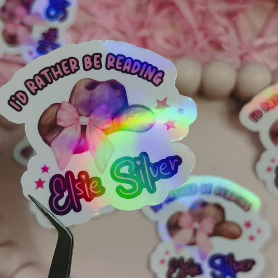 Holographic sticker with a pink-bowed cowboy hat and text reading "I'd Rather Be Reading Elsie Silver" on a bookish background with pink shredded paper