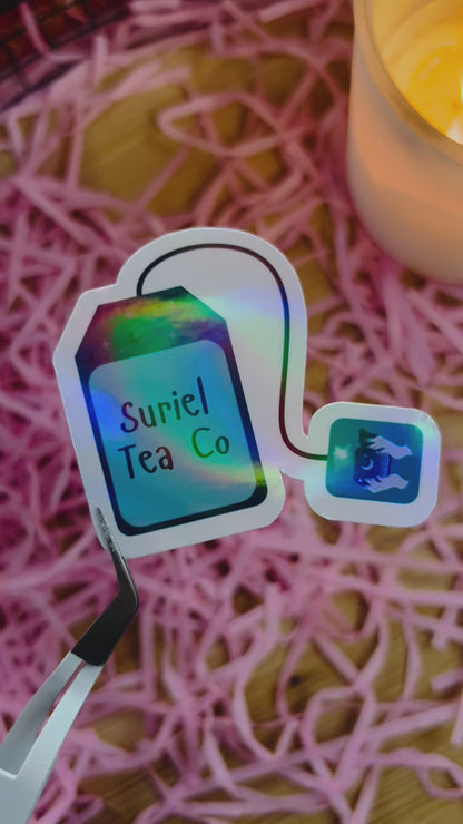 Holographic sticker shaped like a tea bag with the words "Suriel Tea Co," featuring a mystical eye design on the tag, held by tweezers against a pink shredded paper background.