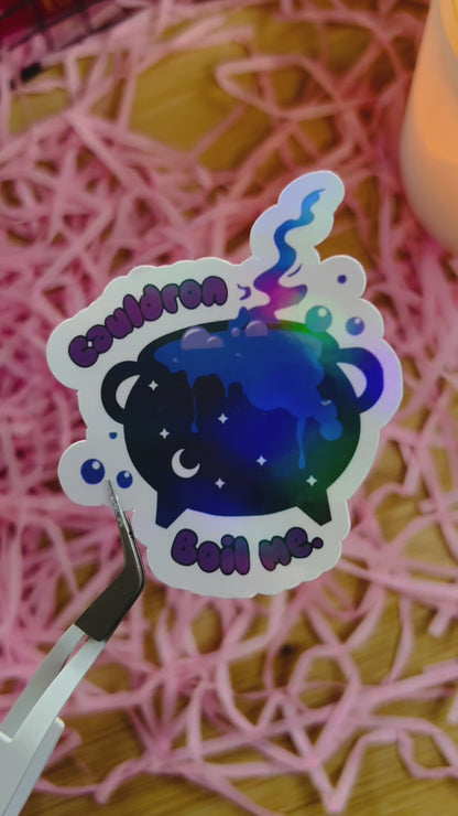 A holographic sticker featuring a celestial cauldron bubbling with magic, inspired by A Court of Thorns and Roses. The words "Cauldron Boil Me" appear in a whimsical font. The sticker is held by tweezers against a pink shredded paper background, with a candle in the corner. 