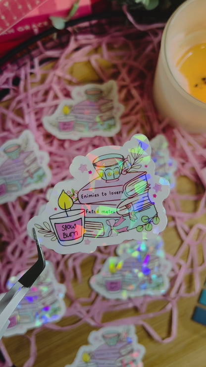 Holographic book stack sticker featuring a tea cup, books labelled with romance tropes (Enemies to Lovers, Slow Burn, Fated Mates), and cosy bookish details.