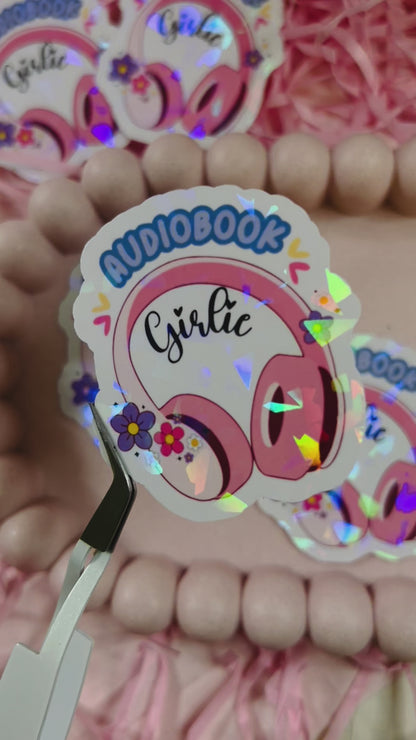 Holographic “Audiobook Girlie” sticker featuring pink headphones, colorful flowers, and sparkly finish — displayed with pastel decor and pink shredded paper.