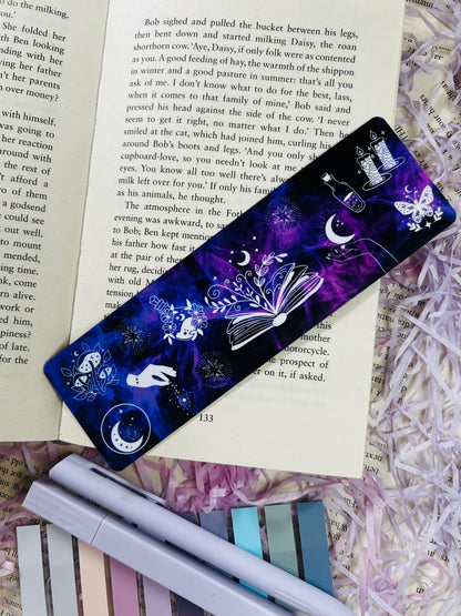 Dark purple witchy bookmark with celestial and magical illustrations including moons, candles, books, and stars, placed on an open book