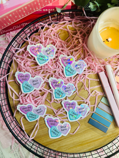 Holographic book trope sticker featuring pastel candy hearts with the phrases "Fated Mates," "Slow Burn," and "Who Did This?" held by tweezers over pink shredded paper.