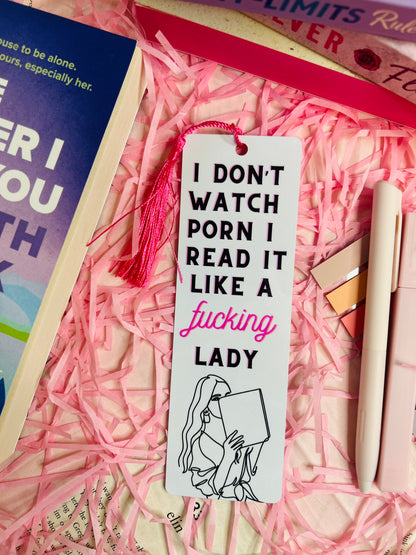 A sassy tassel bookmark with the quote 'I Don’t Watch, I Read It Like a F***ing Lady,' placed on a romance book, surrounded by pink shredded paper, pens, and other books