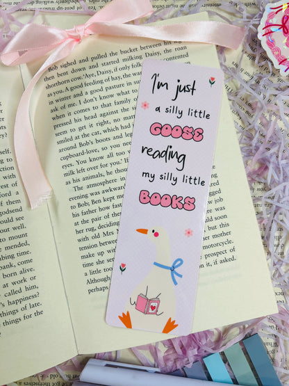 Set of three pastel bookmarks with silly goose and bow designs, placed on an open book with soft pink ribbon, shredded filler, and pens.