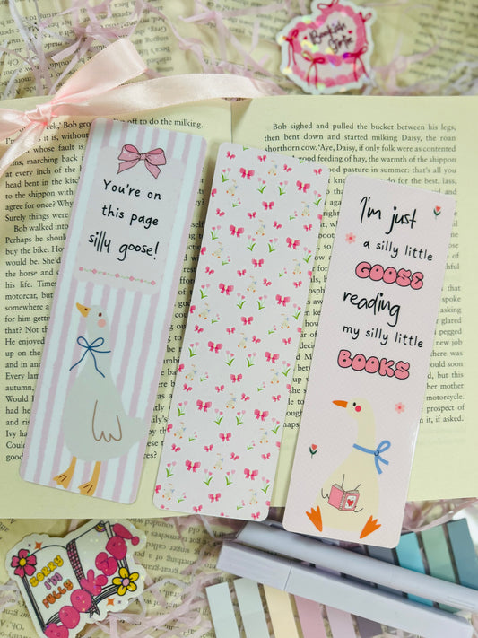 Set of three pastel bookmarks with silly goose and bow designs, placed on an open book with soft pink ribbon, shredded filler, and pens.