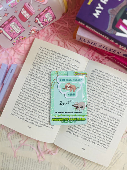 Sloth magnetic bookmark that says ‘You Fell Asleep Here’ placed on an open book with bookish stickers, pens, and novels in the background.