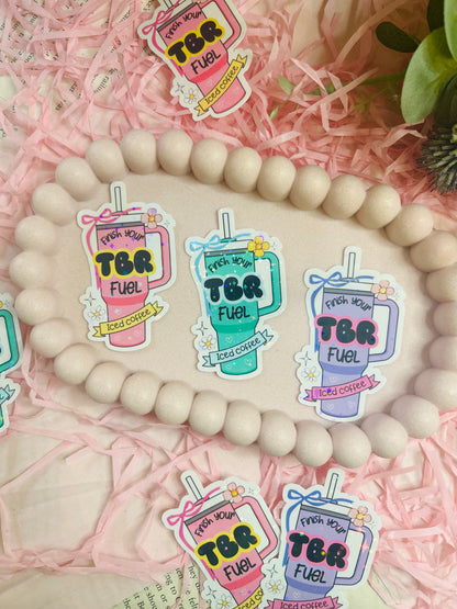 Pastel iced coffee bookish vinyl stickers with "Finish Your TBR Fuel" text, surrounded by pink shredded paper and decorative beads – perfect for readers and book lovers.