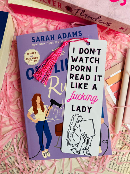 A sassy tassel bookmark with the quote 'I Don’t Watch, I Read It Like a F***ing Lady,' placed on a romance book, surrounded by pink shredded paper, pens, and other books